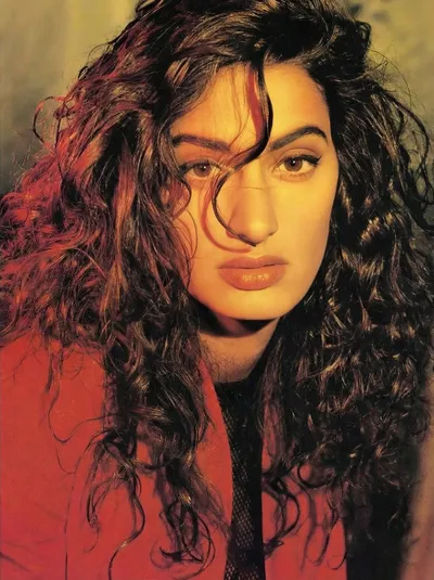 A close-up portrait photo of a young woman with long, voluminous dark curly hair with reddish-brown highlights, wearing a red top or jacket. She has brown eyes, full lips with orange-toned lipstick, and strands of hair falling across her face. The photo has a warm golden-yellow toned background and appears to be a professional editorial or magazine portrait, likely from the 1980s or early 1990s based on the styling and photo quality.