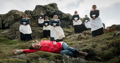 A dramatic scene, likely a film or theater production still, set on a rocky hillside: a woman in a red jacket and jeans lies motionless on the ground with small white flowers placed on her, while seven women dressed in dark period clothing with white aprons and bonnets stand above her looking down.