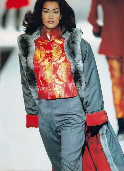 A runway photo of a woman with long dark hair wearing a red silk brocade jacket with gold dragon and floral embroidery, styled over a grey pinstripe trouser. Over this she wears a grey satin coat with a grey fur lining and red satin cuffs and lining. She holds the coat open as she walks the runway.