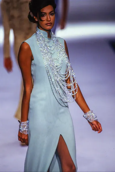 A runway photograph of a woman with dark hair swept up, wearing a sleeveless pale blue column dress with a mandarin collar and a front slit. Draped across the front of the dress are multiple strands of crystal or rhinestone chains that cascade from the neckline diagonally across the bodice. She also wears matching crystal or rhinestone wide cuff bracelets on both wrists.