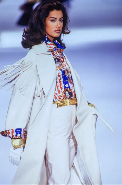 A runway photo of a woman with voluminous dark brown hair, wearing a white fringe leather coat over an Americana-print blouse featuring red, white, blue, and gold stars-and-stripes patterns with what appears to be portrait imagery. She is also wearing white trousers, white gloves with gold cuffs, and a wide gold metallic belt. The coat has long white fringe along the sleeves and shoulders.