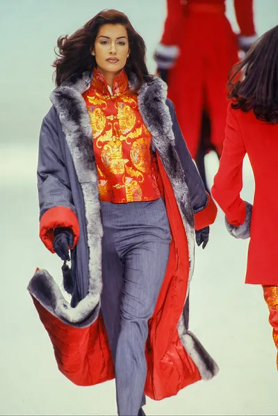 A runway photo showing a woman with long dark hair walking toward the camera. She wears a red-lined, floor-length navy blue coat with grey fur trim, a red mandarin-collar jacket with gold and multicolour brocade pattern, grey straight-leg trousers, and red gloves with black detailing. A second model in a red coat is partially visible to the right, and a third is visible in the background.