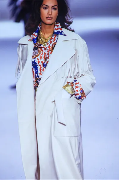 A runway photo of a woman with long dark hair wearing a large oversized white coat with fringe trim across the shoulders, layered over a red, white, and blue printed shirt or blouse with a bold graphic pattern. She accessorizes with layered gold chain necklaces and holds a small gold clutch bag.