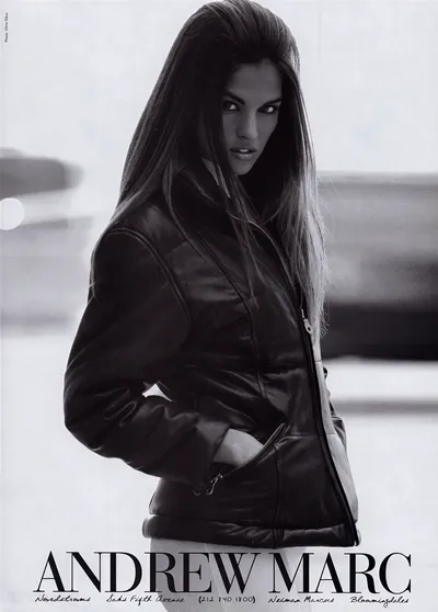 This is a black and white fashion advertisement for Andrew Marc. A woman with long dark hair is shown from the waist up, wearing a leather zip-up jacket with zippered pockets. She is looking back over her shoulder toward the camera. The ad lists retail stockists including Nordstroms, Saks Fifth Avenue, Neiman Marcus, and Bloomingdales, along with a phone number.