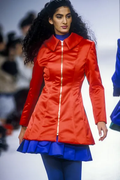 A runway photo of a woman with long dark curly hair walking a fashion show catwalk. She wears a fitted red satin zip-front coat dress with a gold zipper running from hem to collar, a wide pointed collar, and a flared skirt hem trimmed with a blue satin ruffle. Beneath the coat she wears a cobalt blue turtleneck top and cobalt blue tights or trousers.
