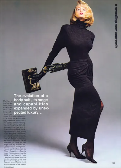 This is a fashion magazine spread (page 133) featuring a woman with short blonde hair wearing a dark charcoal grey long-sleeve turtleneck bodysuit and a separate dark charcoal grey sarong-style midi skirt, paired with black leather elbow-length gloves, black high-heel pointed-toe pumps, and holding a black and gold textured clutch bag. The text describes the clothing as a Donna Karan New York cashmere bodysuit and matching skirt, with accessories including Wendy Gell cuffs, Carlos Falchi gloves, and Hanes pantyhose. The photo credit reads Andrea Blanch.