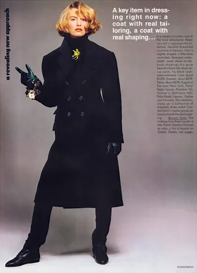 A magazine fashion spread featuring a woman with short blonde hair wearing a long black double-breasted cashmere coat over a black turtleneck and black stirrup pants, paired with dark reptile-skin gloves, multiple bracelets, and black lace-up ankle boots. A small yellow floral brooch is pinned at the coat's neckline. The photographer credit "Andrea Blanch" appears in the bottom right corner.