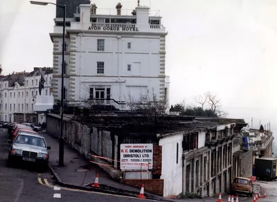 A historical photograph showing the Avon Gorge Hotel, a large white multi-storey building in Bristol, England. In the foreground is a demolition site with signage for "R.C. Demolition (Bristol) Ltd." with phone numbers 0272 231320/231321, orange traffic cones, safety tape, and several parked cars from what appears to be the late 1980s based on the vehicle registration plates.