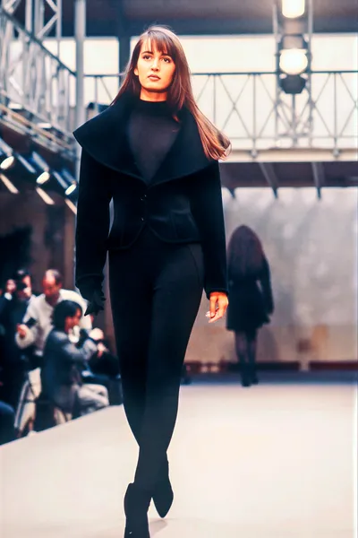 A runway photo of a woman with long brown hair and bangs wearing an all-black outfit consisting of a wide-lapel structured jacket, slim trousers, and pointed boots, walking toward the camera on a white catwalk with seated audience members visible to the left and another model in the background.