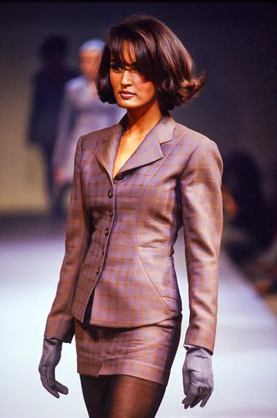 A runway photo of a woman with short, voluminous dark hair walking on a white catwalk, wearing a fitted plaid blazer and matching mini skirt in grey and orange tones, paired with long grey gloves and black tights.