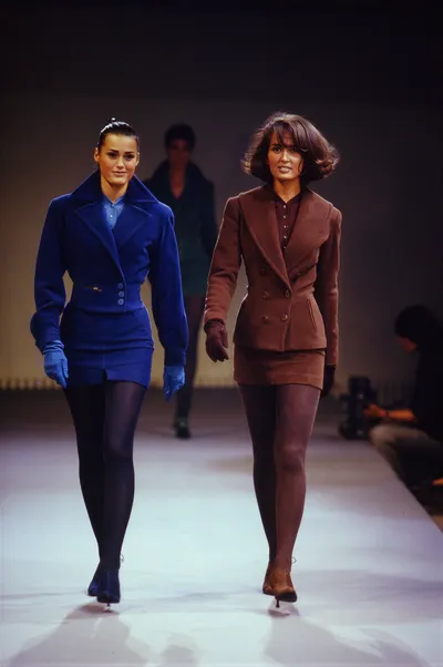 A runway photo showing two women walking side by side on a white catwalk. The woman on the left wears a cobalt blue structured jacket, matching mini skirt, blue gloves, and dark tights; the woman on the right wears a brown double-breasted blazer, matching mini skirt, dark gloves, and brown tights. A third model is visible in the background.