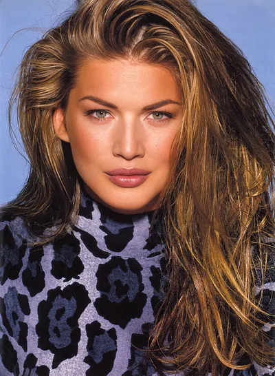 A close-up portrait photograph of a woman with long highlighted brown hair, green eyes, and a tanned complexion, wearing a blue and black leopard-print turtleneck top, photographed against a light blue background. The image has the look of a professional editorial or magazine portrait from the late 1980s or early 1990s.
