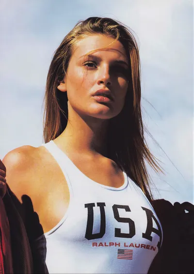 A fashion advertisement showing a young woman with long brown hair wearing a white Ralph Lauren "USA" tank top, photographed from below against a partly cloudy sky.