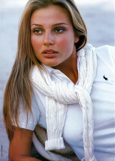 A fashion advertisement showing a young woman with long straight brown hair wearing a white Polo Ralph Lauren polo shirt and a white cable-knit scarf loosely tied around her neck. A phone number (1-877-276-7656) is printed vertically along the left edge of the image.