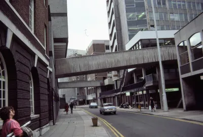A street-level photograph of an urban area, likely a British city from the 1980s, showing a road with double yellow lines, concrete brutalist pedestrian walkways and bridges spanning between buildings, parked and moving cars, and several pedestrians on the pavement. On the left, a woman with dark curly hair wearing a pink top is pushing a pram, and a dark brick building with arched windows is visible alongside modern concrete office blocks.