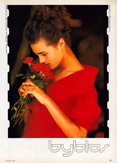 This is a vintage fashion advertisement from Vogue, April 1988, for the brand Byblos. It shows a woman with dark curly hair pinned up, wearing a red structured dress with angular shoulders, holding a bouquet of red roses close to her face as she smells them. The brand name Byblos appears in outline lettering at the bottom of the page, and the credits identify the art director as Nando Miglio and photographer as Eddy Kohli.