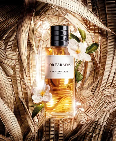 A product advertisement showing a glass bottle of "Dior Paradise" eau de parfum by Christian Dior Paris, lying on its side surrounded by white flowers with green leaves, set against a golden woven palm leaf backdrop.