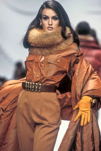 A runway photo of a woman with long dark hair wearing a voluminous copper/rust-toned iridescent coat with a large fur collar, gold buttons, a wide dark belt with gold chain detailing, brown wide-leg trousers, and mustard-yellow leather gloves.