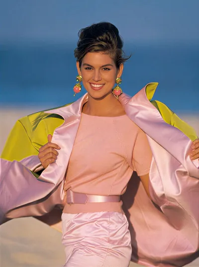 A fashion photograph, likely from the 1980s, showing a smiling woman with short dark hair wearing a pink knit top, pink satin skirt, pink belt, and large gold-and-pink drop earrings, holding open a yellow-and-pink satin jacket, with a blurred beach and sky in the background.