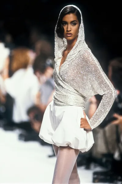 A runway photo of a woman with dark hair wearing a silver metallic hooded mesh top and a white bubble-hem mini skirt with white tights, posed with one hand on her hip on a catwalk with a blurred audience in the background.