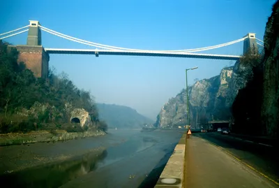 A photograph taken from road level beneath the Clifton Suspension Bridge, showing the bridge spanning a rocky gorge above the River Avon at low tide, with muddy riverbanks exposed, steep limestone cliffs on both sides, and a road with a concrete barrier running along the right side.