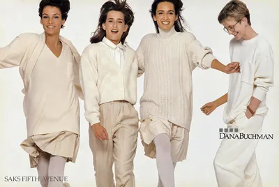 This is a vintage fashion advertisement for Dana Buchman at Saks Fifth Avenue, showing four women posed together against a white background, all dressed in coordinating cream and ivory knitwear. From left to right: a woman with short dark hair wearing a cream V-neck sweater over a beige skirt with white tights; a young woman with long dark hair wearing a cream checkered cardigan over a white collared shirt and beige trousers; a woman with dark hair wearing a chunky cream cable-knit oversized sweater over a beige skirt with white tights; and a woman with short light hair wearing glasses and a plain cream sweatshirt with cream trousers and a brown wristwatch.