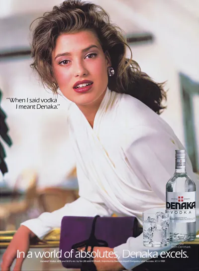 A vintage 1989 print advertisement for Denaka Vodka featuring a woman with voluminous dark brown hair wearing a white draped top, leaning forward toward the camera. A bottle of Denaka Vodka and two glasses of ice are shown in the lower right corner, with the tagline "In a world of absolutes, Denaka excels."