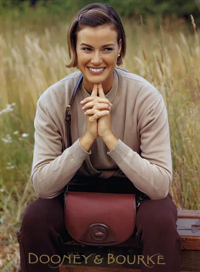 A Dooney & Bourke advertisement featuring a smiling woman with short brown hair, wearing a light beige ribbed sweater and dark burgundy trousers, seated outdoors on what appears to be a vintage trunk. She holds a burgundy pebbled-leather Dooney & Bourke crossbody saddle bag on her lap, with tall dry grass visible in the background.