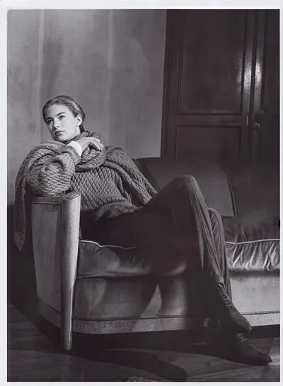 Black-and-white fashion advertisement or editorial photograph showing a woman with slicked-back hair seated in a large upholstered armchair. She wears an oversized, heavily cable-knit sweater with exaggerated rope-like textures, slim trousers, and lace-up ankle boots; a wooden-panelled door is visible in the background.