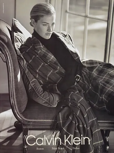 A black-and-white Calvin Klein print advertisement featuring a woman with slicked-back blonde hair, seated in a wooden armchair, wearing a dark turtleneck top and a large plaid coat or wrap draped loosely around her body; the ad lists the store locations Boston, Palm Beach, and Dallas at the bottom.