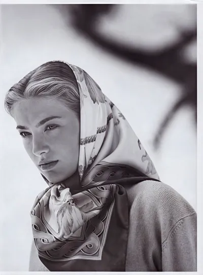 Black-and-white fashion portrait of a young woman with slicked-back light hair, wearing a printed silk scarf tied over her head and knotted at the neck, featuring a floral and ornamental pattern with what appears to be an animal motif at the front. She wears a light-colored knit sweater underneath. The background is pale and out of focus, with a dark blurred shape visible in the upper right corner.