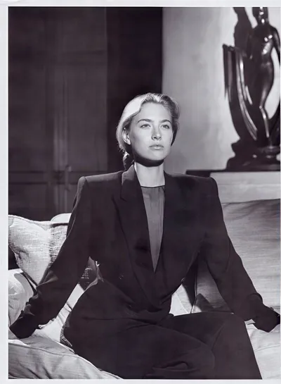 A black-and-white portrait photograph of a woman with light blonde hair pulled back, seated on a sofa with cushions. She is wearing a dark structured blazer with pronounced shoulders over what appears to be a dark top or dress beneath it; whether the blazer and bottom constitute a matching set is unclear due to the monochrome photography. In the background is a decorative sculptural figure on a surface, and dark wood paneling.