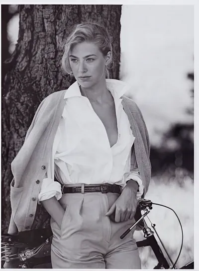 A black-and-white fashion portrait of a woman with blonde hair pulled loosely up, leaning against a tree trunk with a bicycle partially visible beside her. She wears an open-collared white button-front shirt with sleeves rolled up, light-coloured high-waisted trousers with a dark belt, and a light-coloured cardigan draped over her shoulders.