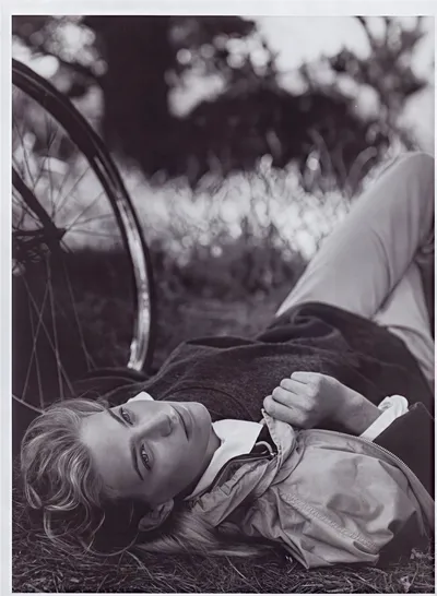 Black-and-white editorial or magazine photograph of a young woman with light, loosely styled hair, lying on her back in grass next to a bicycle wheel. She is wearing a zip-up jacket over a collared shirt and light-coloured trousers, and she is looking upward toward the camera with a relaxed expression.