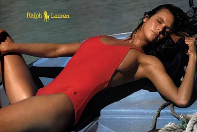 A Ralph Lauren advertisement showing a woman with dark wet hair reclining on a boat, wearing a red one-piece swimsuit with a small polo logo on the hip, with water visible in the background.