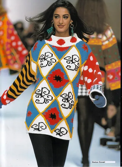 A runway photo of a woman with long dark hair wearing an oversized knit sweater in a diamond/argyle pattern featuring blue, white, red, and yellow sections decorated with floral motifs and fleur-de-lis designs; the sleeves are mismatched, one with yellow and black stripes and one with a red polka-dot pattern. She is paired with black leggings and teal drop earrings. The designer label "Enrico Coveri" appears in the bottom right corner.
