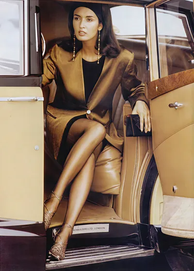 A fashion advertisement photograph showing a woman with long dark hair wearing a gold/bronze blazer and skirt suit, gold drop earrings, and strappy heeled sandals, seated in the open doorway of a vintage luxury car with cream-colored leather interior; a small placard on the car's step reads "Coachbuilders Ltd. London."