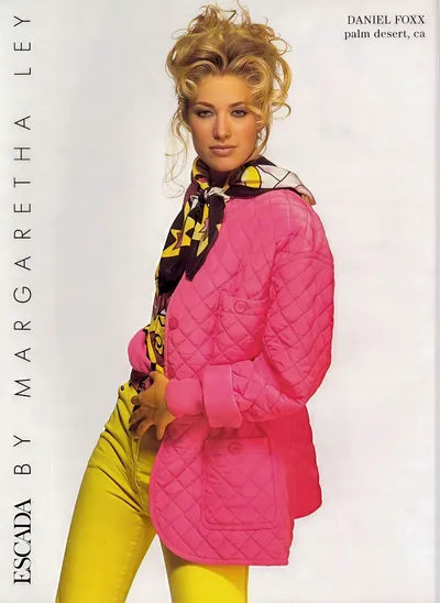 A vintage fashion advertisement for Escada by Margaretha Ley, sold at Daniel Foxx in Palm Desert, CA. A woman with blonde upswept curly hair poses wearing a bright pink quilted jacket, yellow fitted pants, and a multicolored printed silk scarf tied loosely at the neck in yellow, black, white, and brown tones.