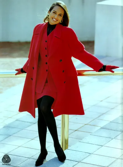 This is a fashion advertisement featuring the Pure New Wool certification logo in the bottom left corner. A woman with long brown hair is posing outdoors on a tiled surface, leaning against a gold railing. She is wearing a red double-breasted overcoat open over a red buttoned vest and a red skirt, paired with a black turtleneck, black tights, black heeled shoes, and red gloves with black trim at the wrist. She is also wearing large gold geometric earrings and is smiling at the camera.