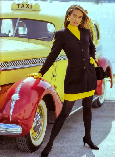 This appears to be a fashion magazine spread or catalog advertisement from what looks like the late 1980s or early 1990s. A woman with long brown hair poses leaning against the red front fender of a vintage yellow taxi cab. She wears a black long cardigan jacket over a yellow turtleneck sweater, a yellow mini skirt with a black hem band, black tights, black heeled pumps, yellow leather gloves, and gold square earrings, and she carries a small black handbag. The taxi has a checkered stripe along its body and a rooftop taxi sign.