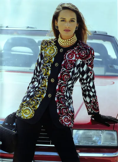 This appears to be a fashion magazine editorial or advertisement photograph from the early 1990s, featuring a woman with long brown hair leaning against the hood of a red car. She is wearing a boldly patterned blazer jacket that features a harlequin diamond check pattern in black and white combined with baroque-style ornamental motifs in gold on the left side and deep red and burgundy on the right side, with gold logo buttons down the center front. She is accessorized with a multi-strand gold pearl choker necklace, large gold drop earrings, and black gloves, and is wearing black trousers underneath the jacket.