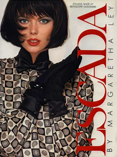 A vintage fashion advertisement for Escada by Margaretha Ley, available at the Escada Shop at Bergdorf Goodman. A woman with a short dark bob haircut, blue eyes, and red lipstick wears a black-and-white geometric and paisley-patterned blouse with a black satin bow at the neck and black suede gloves. The brand name "ESCADA" is printed vertically in large red letters on the right side of the image.