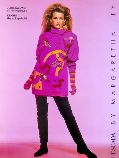 A vintage fashion advertisement for Escada by Margaretha Ley, featuring a woman with voluminous auburn hair wearing a bright purple oversized turtleneck sweater printed with abstract shapes and embellished with sequined shoe motifs in pink and gold, paired with striped orange-and-purple long gloves, black fitted pants, and tall black boots. The background is a gradient pink-purple, and text in the upper left lists retail locations in St. Petersburg, FL and Grand Rapids, MI.
