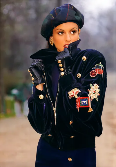 A fashion photograph, likely a magazine editorial or advertisement, showing a woman with brown eyes and red lipstick wearing a dark navy blue velvet zip-up bomber jacket heavily embellished with gold buttons and colorful beaded patches depicting motifs such as a carriage, a medal/star, and a flag. She is also wearing a dark green and navy plaid beret, black leather gloves adorned with gold buttons, and gold heart-shaped earrings. The background is an outdoor setting with blurred trees.
