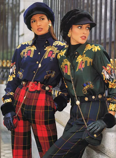 A magazine spread (page 62) showing two women posed against iron railings. The woman on the left wears a navy blue beret, a navy satin blouse printed with gold baroque scrollwork and British royal guard/carriage motifs, red and black tartan wide-leg trousers, a red belt, and navy leather gloves. The woman on the right wears a black fur-trimmed cap, the same blouse style in dark green with identical gold and royal guard print, dark navy and green tartan wide-leg trousers, a dark belt with gold coin-style buttons, and black leather gloves with fur cuffs. Both wear gold heart-shaped drop earrings and carry gold chain-strap bags.