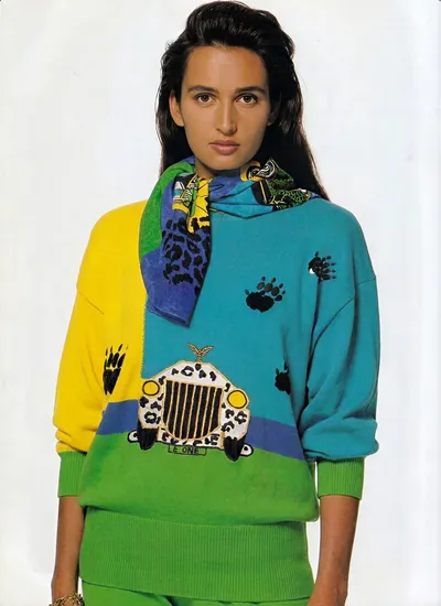 A fashion photograph, likely from a magazine editorial or advertisement from the 1980s, showing a woman with long dark hair wearing a color-blocked knit sweater in yellow, teal/turquoise, blue, and green sections, decorated with black paw print motifs and an embroidered patch depicting a leopard-print jeep with the text "E ONE." She also wears a multicolored silk scarf in blue, green, yellow, and black leopard print loosely draped around her neck, and green knit trousers that appear to match the green section of the sweater.