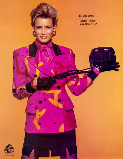 A fashion advertisement for Jacobson's / Daniel Foxx of Palm Desert, CA, featuring a woman with short, voluminous upswept brown hair wearing a bright magenta double-breasted blazer with an abstract geometric pattern in orange, red, and black, black lapels, and black buttons. She has a pink bow-tied scarf at the neck, a black mini skirt, black leather gloves, and gold clip earrings, and is holding a small dark navy velvet hat with gold embellishments. The background is a solid orange gradient. A "Pure New Wool" logo appears in the bottom left corner.