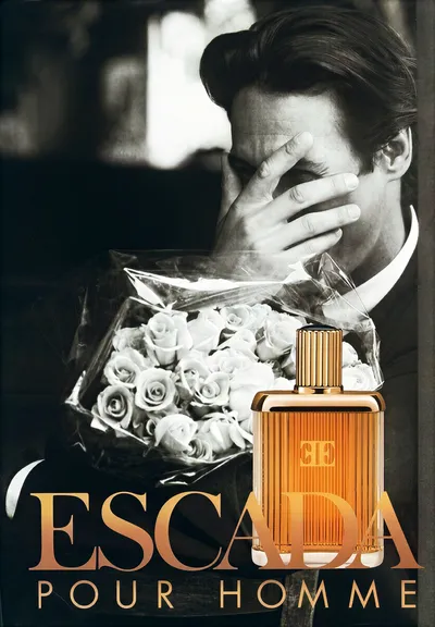 A black-and-white fragrance advertisement for Escada Pour Homme, showing a dark-haired man in a suit holding a bouquet of roses while covering part of his face with one hand; a gold perfume bottle is overlaid in the foreground alongside the brand name in gold text.