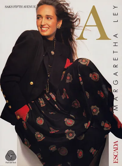 This is a vintage print advertisement for Escada by Margaretha Ley, sold at Saks Fifth Avenue. A woman with long dark brown hair (blowing in the wind) and a bright smile wears a black blazer with gold buttons over a black outfit, along with a voluminous black skirt or wrap covered in a repeating heraldic crest pattern in red and gold; the blazer has red lining visible at the cuffs. The text "Saks Fifth Avenue," "Margaretha Ley," and "Escada" appear on the ad, along with a Pure New Wool certification logo in the bottom left corner.
