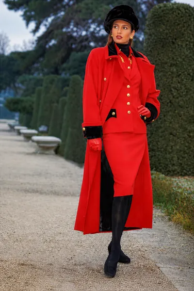 A fashion editorial or advertisement photograph showing a woman with long brown hair walking along a gravel garden path lined with trimmed hedges and stone planters. She wears a long red wool open coat with black velvet cuffs and gold buttons over a red double-breasted jacket with black velvet lapels and gold buttons, a red skirt, a black turtleneck, black tights, black shoes, red leather gloves, a black fur or velvet beret-style hat, and gold earrings.